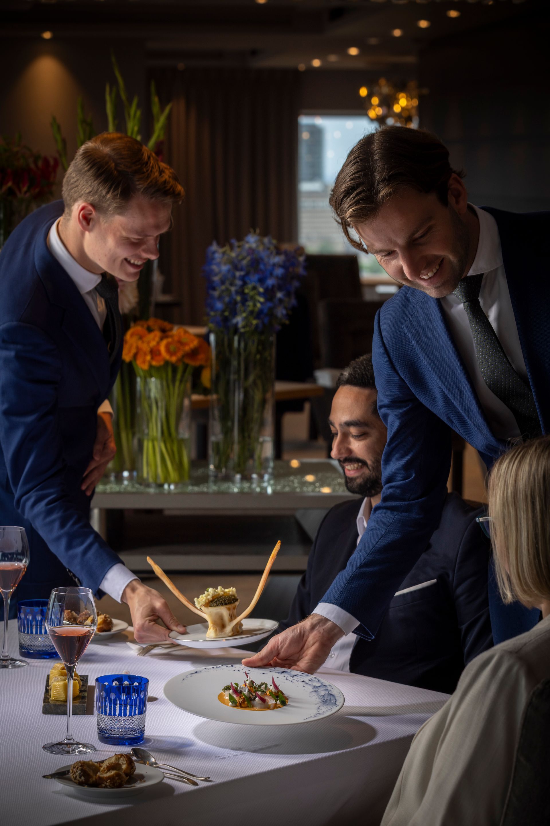 Two waiters serving a dish to a diner in a restaurant with floral arrangements.