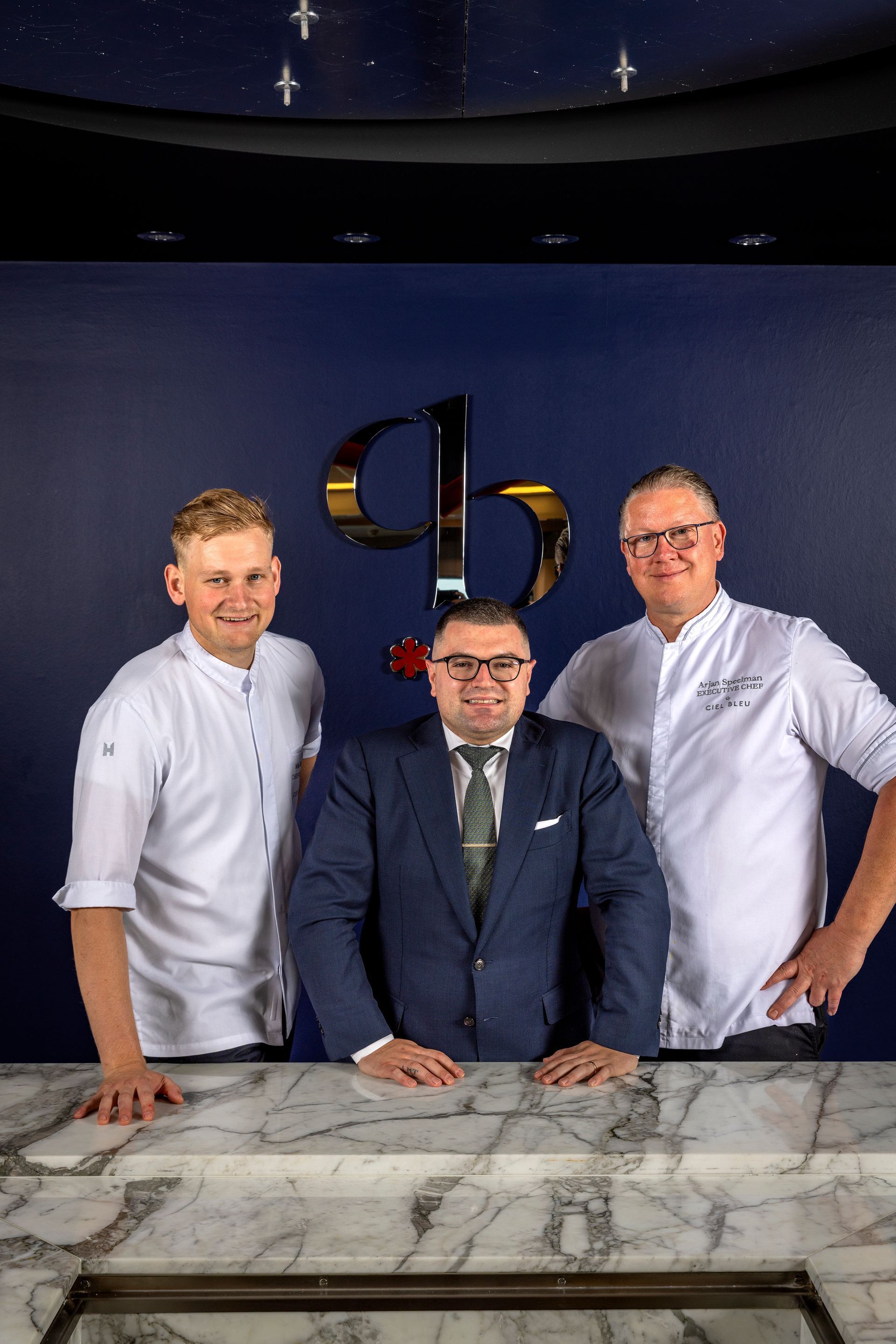 Three people posing in front of a blue wall with a logo. Two in white chef coats, one in a suit.