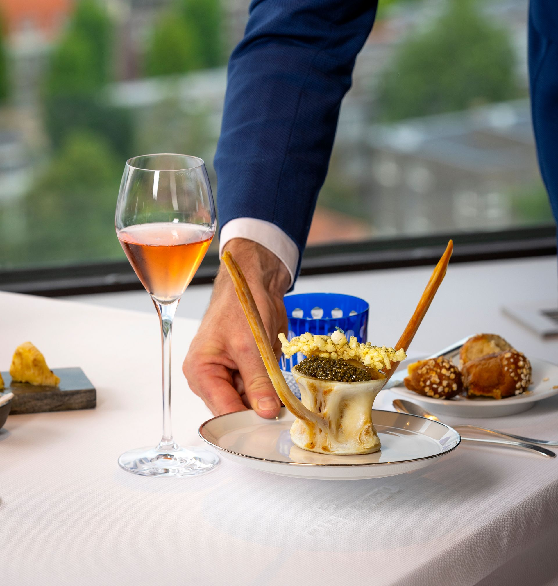 Een ober zet een voorgerecht op een tafel die gedekt is met een glas roséwijn, een wit tafelkleed en uitzicht op de stad.