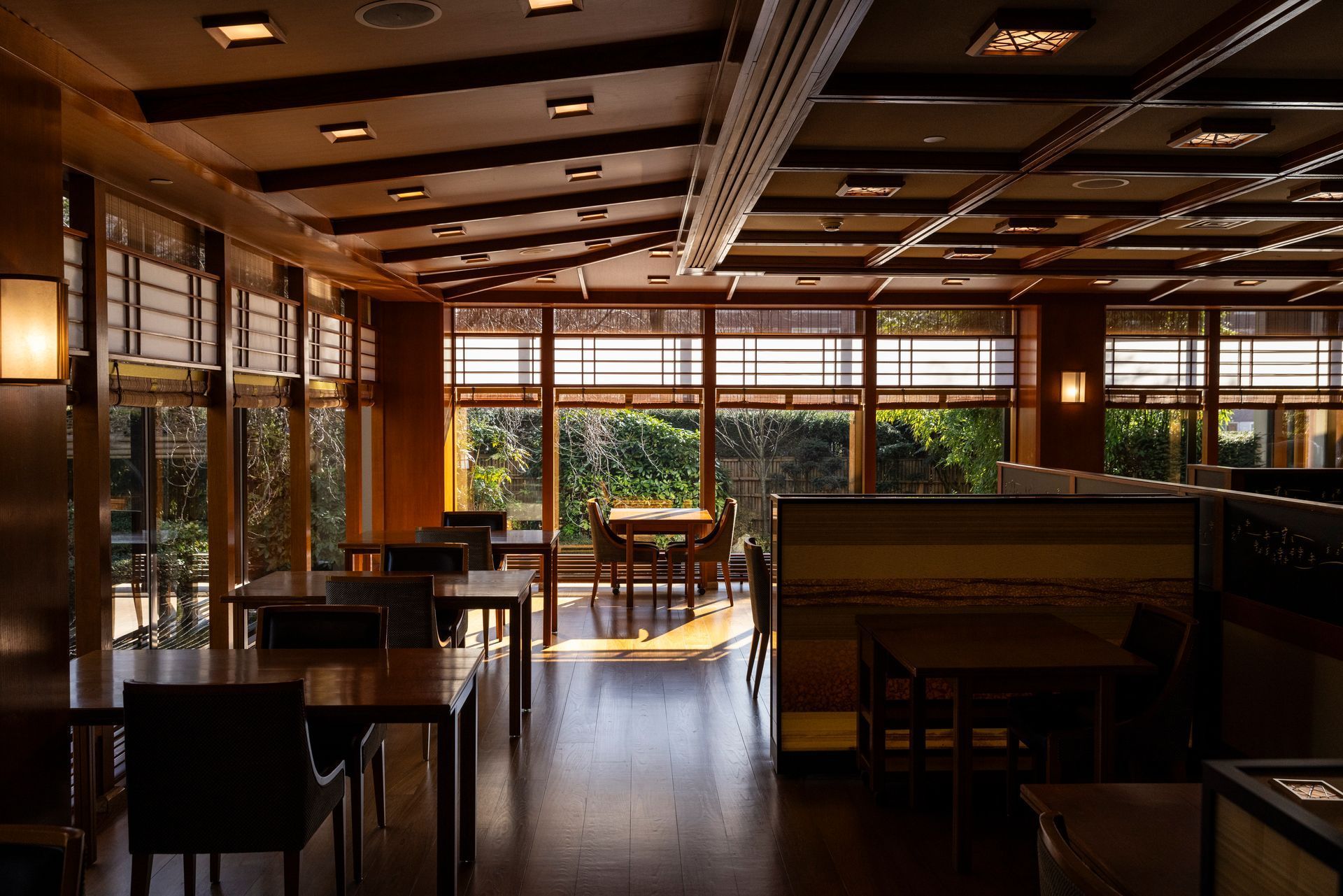 Restaurant interior with wood tables and chairs, large windows overlooking greenery, and warm lighting.