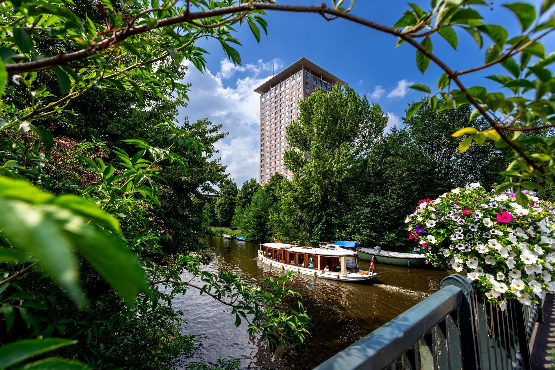 A boat on a river in front of a tall building framed by lush green foliage and flowers.