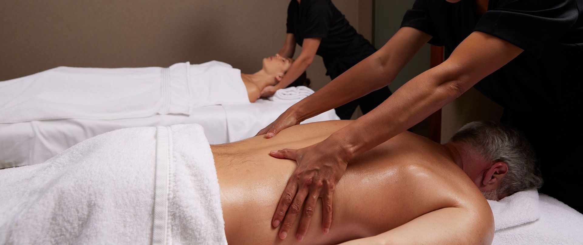 Two people receiving massages in a spa, lying on white sheets. Two massage therapists work simultaneously.
