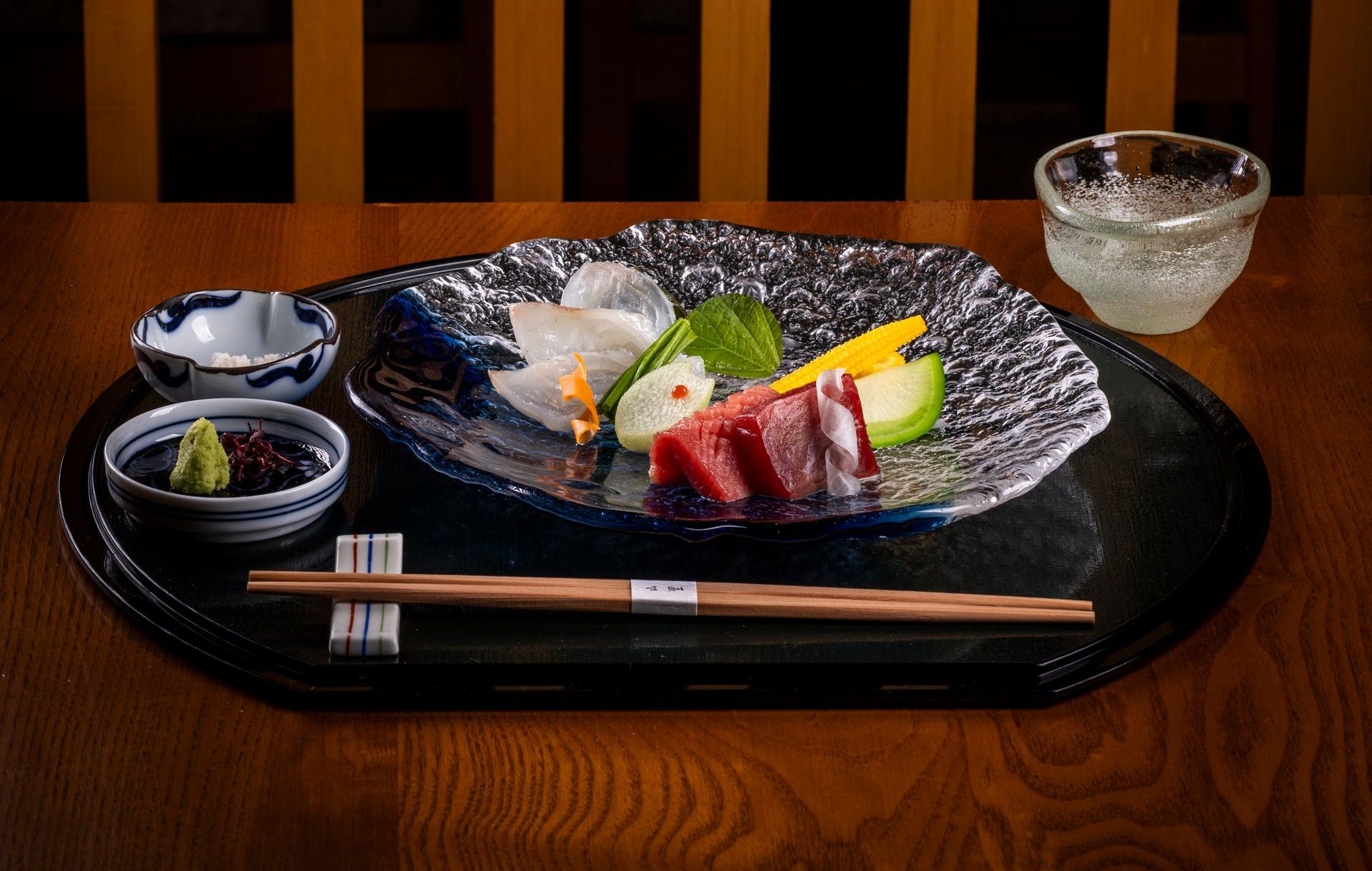 Een bord met sushi en sashimi, eetstokjes, sojasaus, wasabi en een sakeglas op een houten tafel.