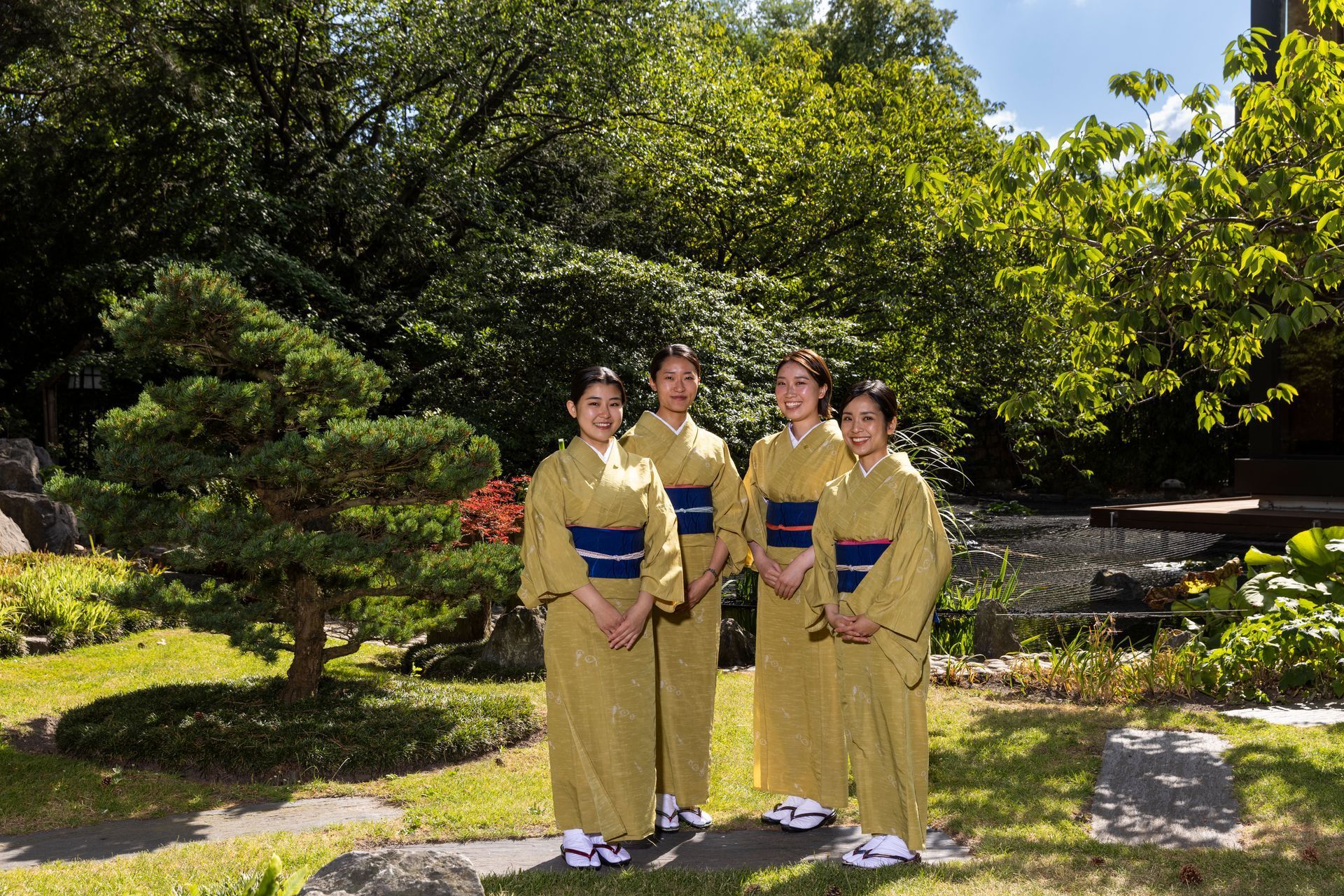 Four people in yellow kimonos stand in a Japanese garden, near a small tree.