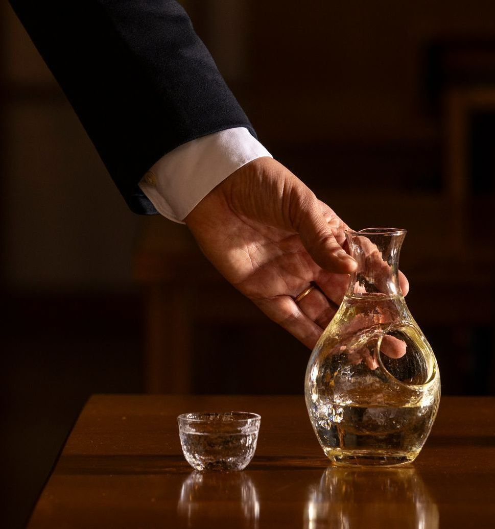 Hand pouring liquid from a carafe into a small glass on a wooden table.