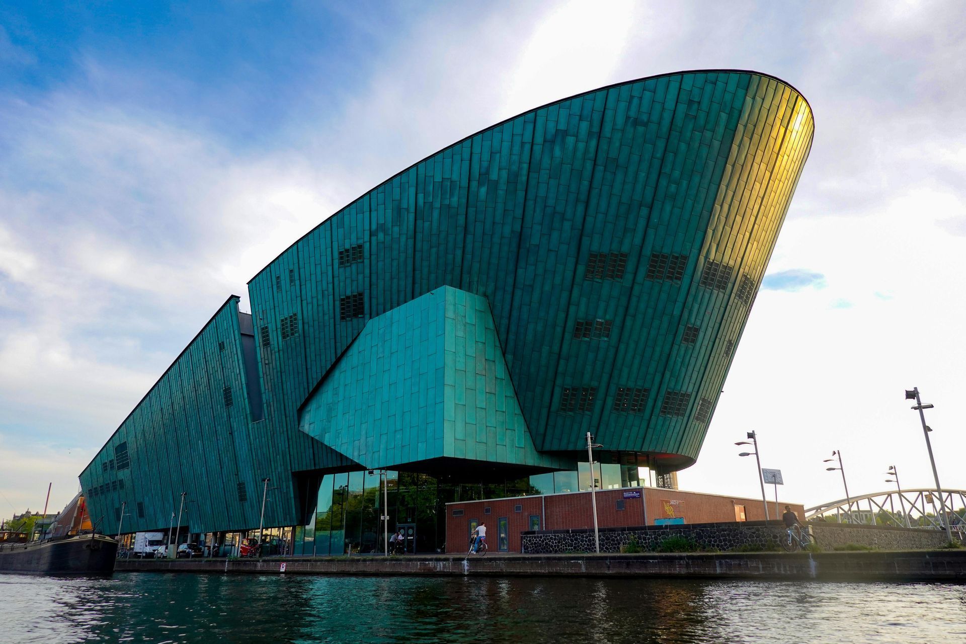 A large, green-paneled science museum building on a waterway; bronze-colored finish with a curved facade.
