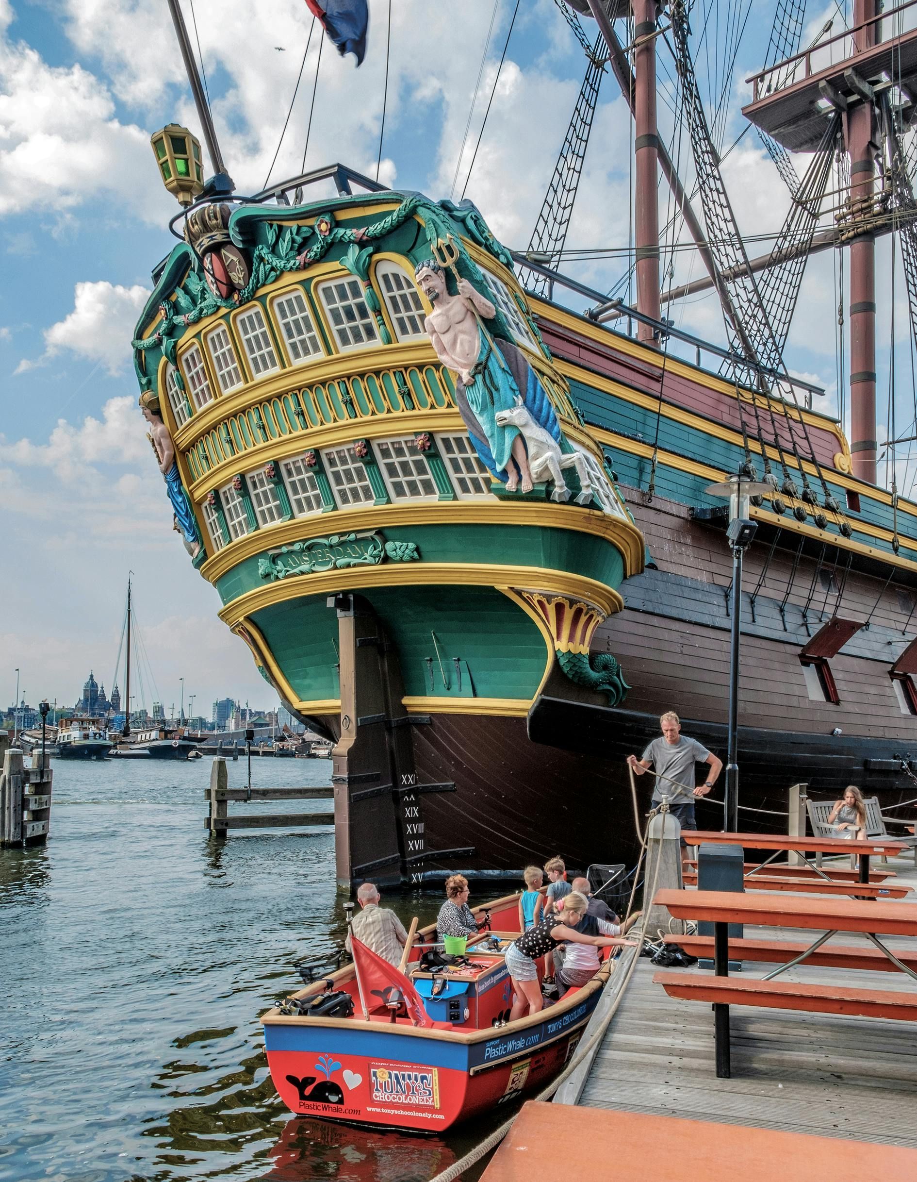 Ornate green and brown ship's stern. Small red boat with people on the water. People on dock.