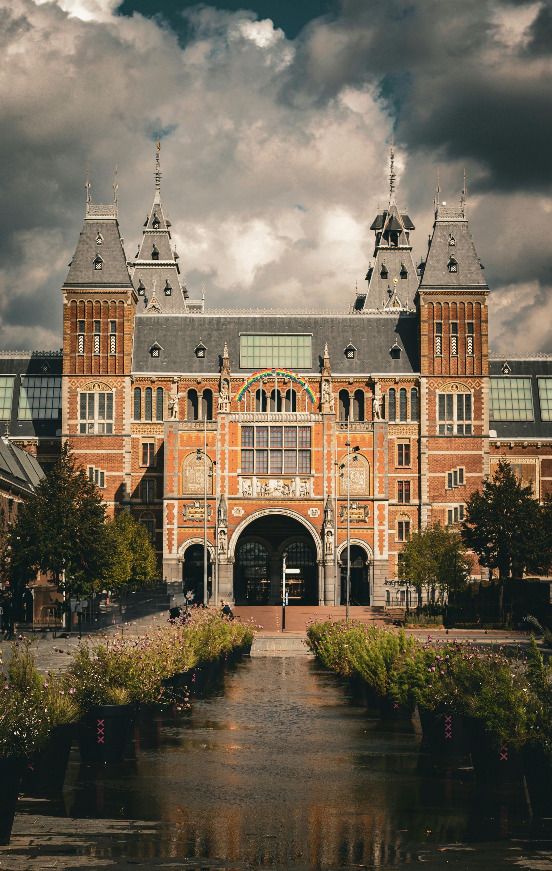 Rijksmuseum, Amsterdam: ornate brick building with towers and arched entrance, cloudy sky overhead, reflecting in a canal.