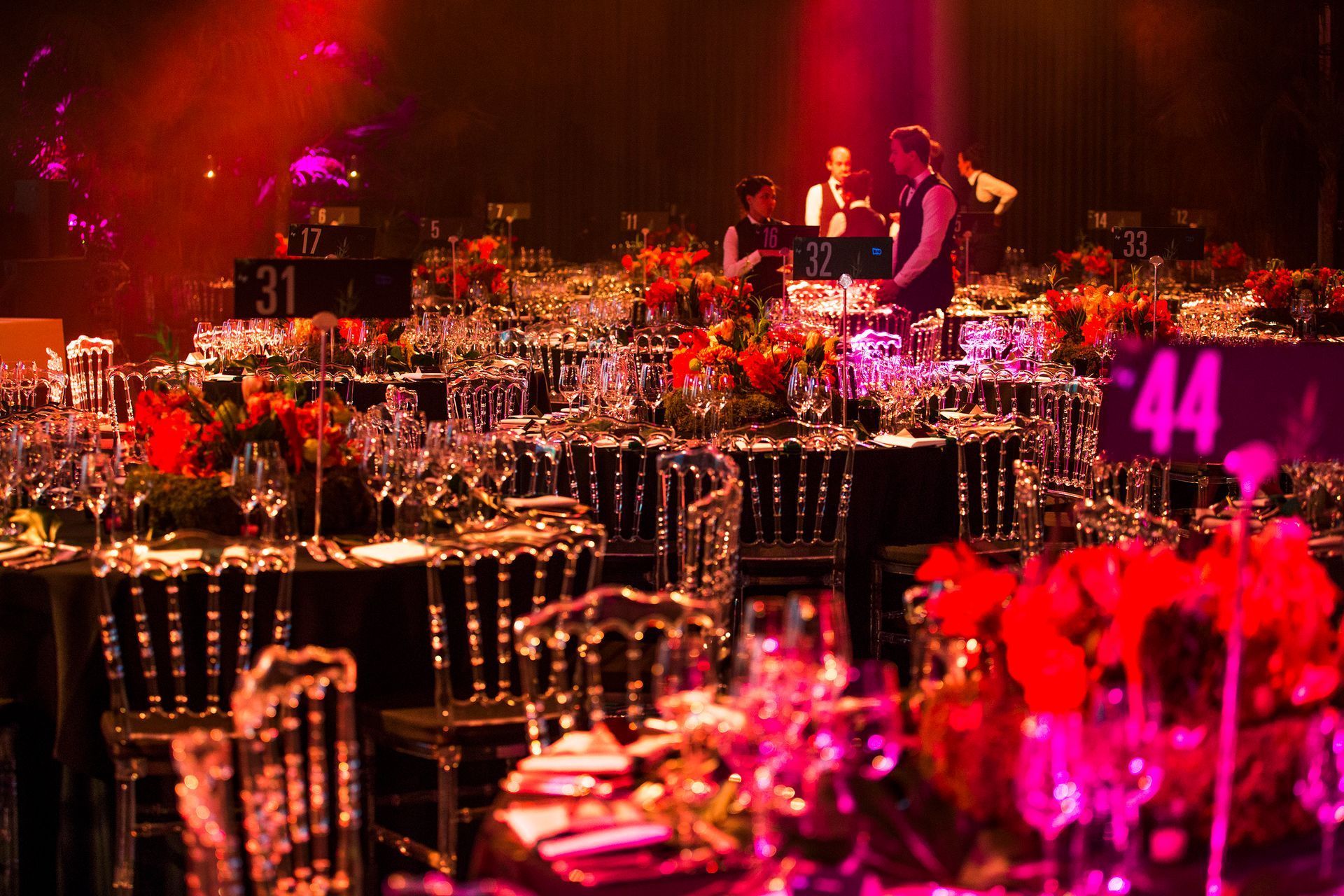 Tables set for a gala dinner, with clear chairs, red flower centerpieces, and staff in the background.