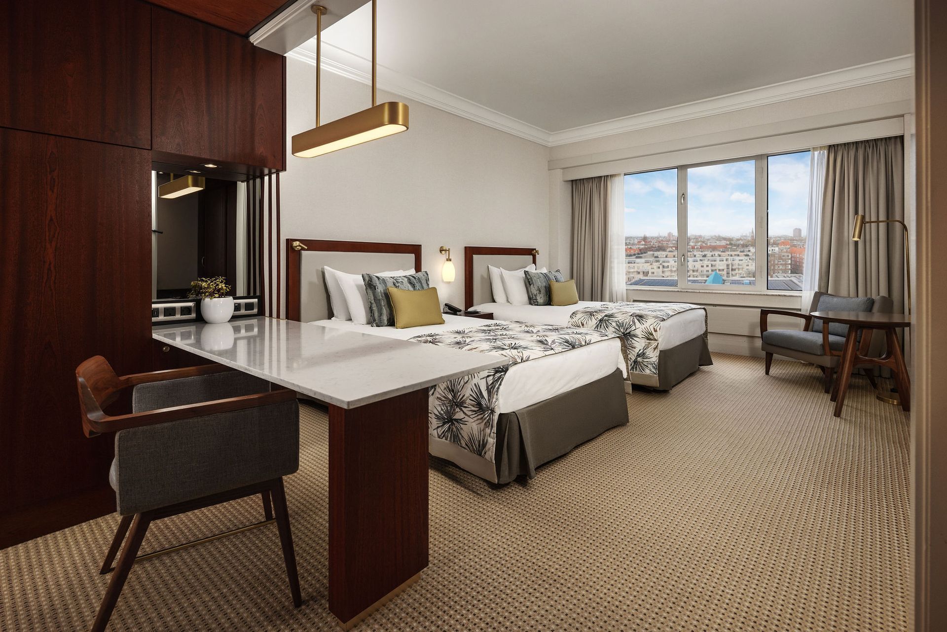 Hotel room with two beds, a desk, and a city view through a large window.