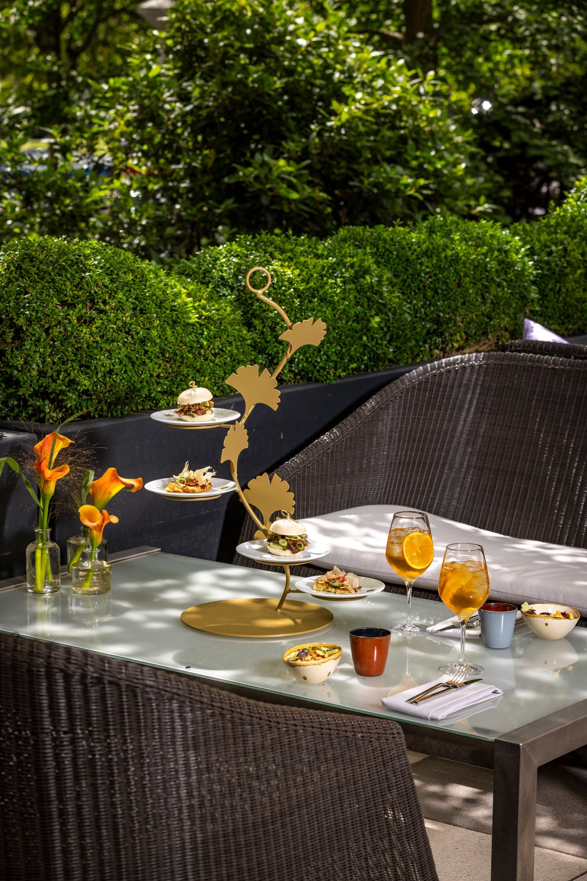 Outdoor table setting with food on a tiered stand, cocktails, and flowers, against a backdrop of greenery.