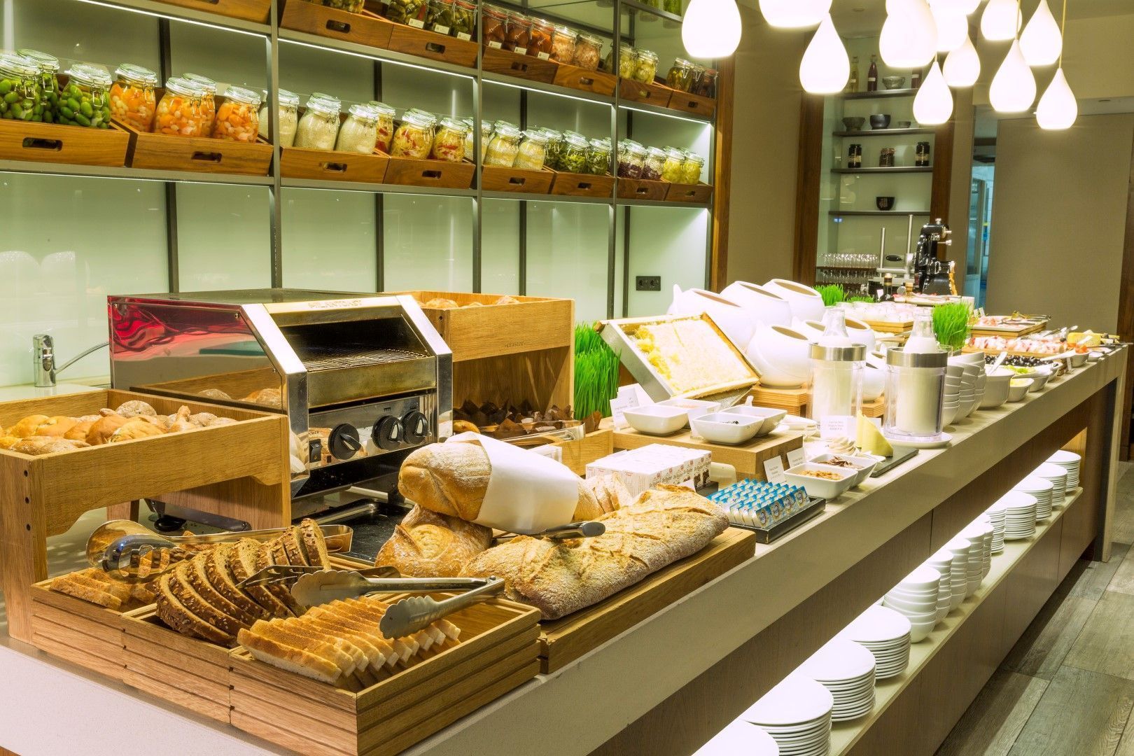 Breakfast buffet with bread, pastries, and condiments. Shelves hold jars of food. Warm lighting.
