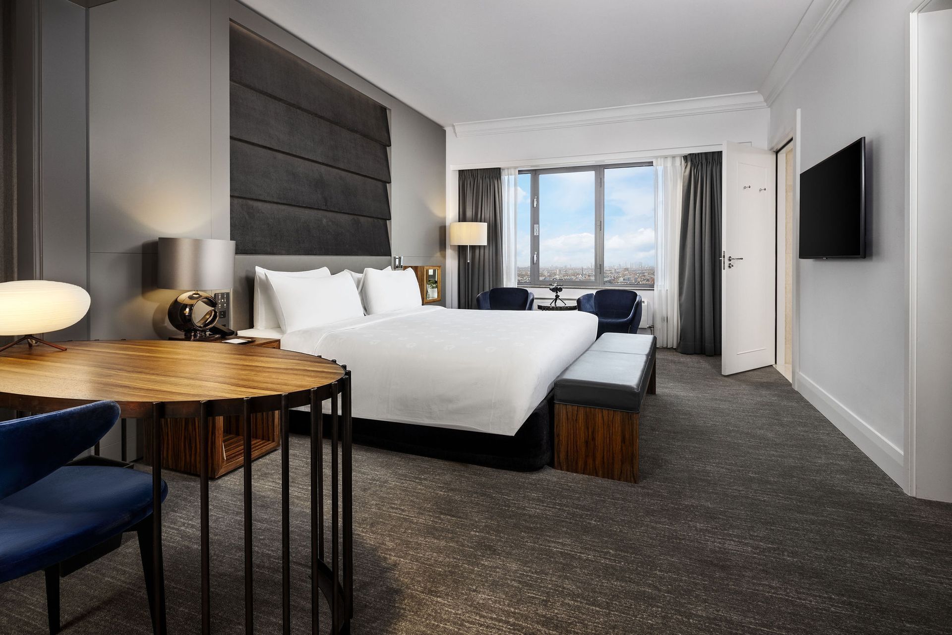 Hotel room with a bed, desk, and a window. Gray walls and carpet with dark blue accents.