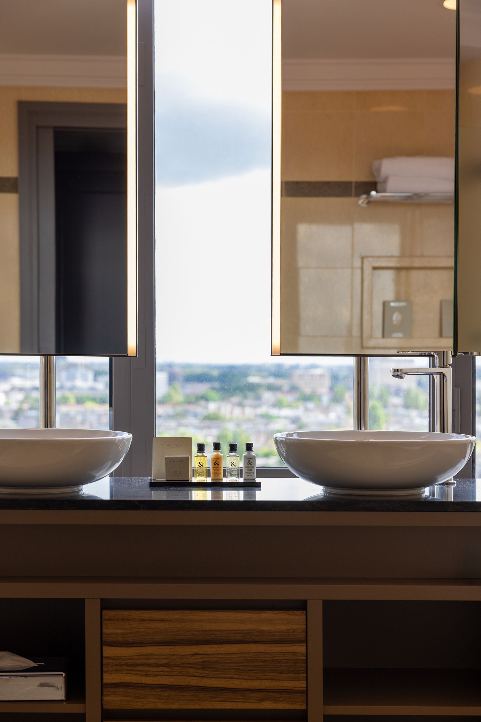 Bathroom with two white sinks, black countertop, tall mirrors, window with city view, and toiletries.