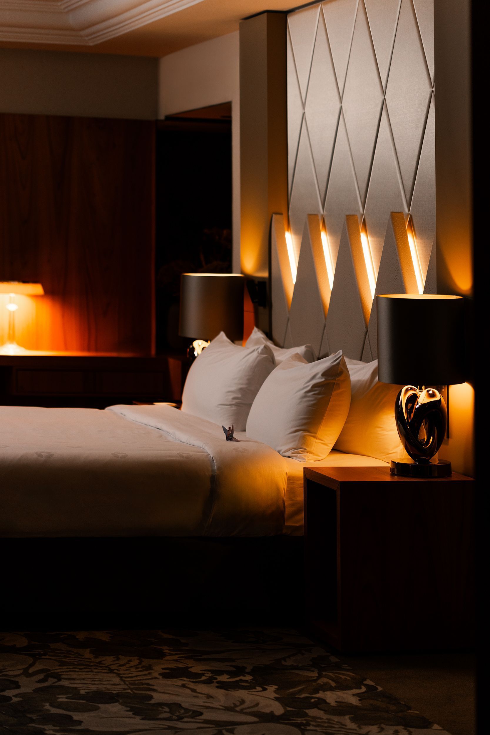 Cozy hotel bedroom with a lit bed, bedside lamps, and a textured wall feature.