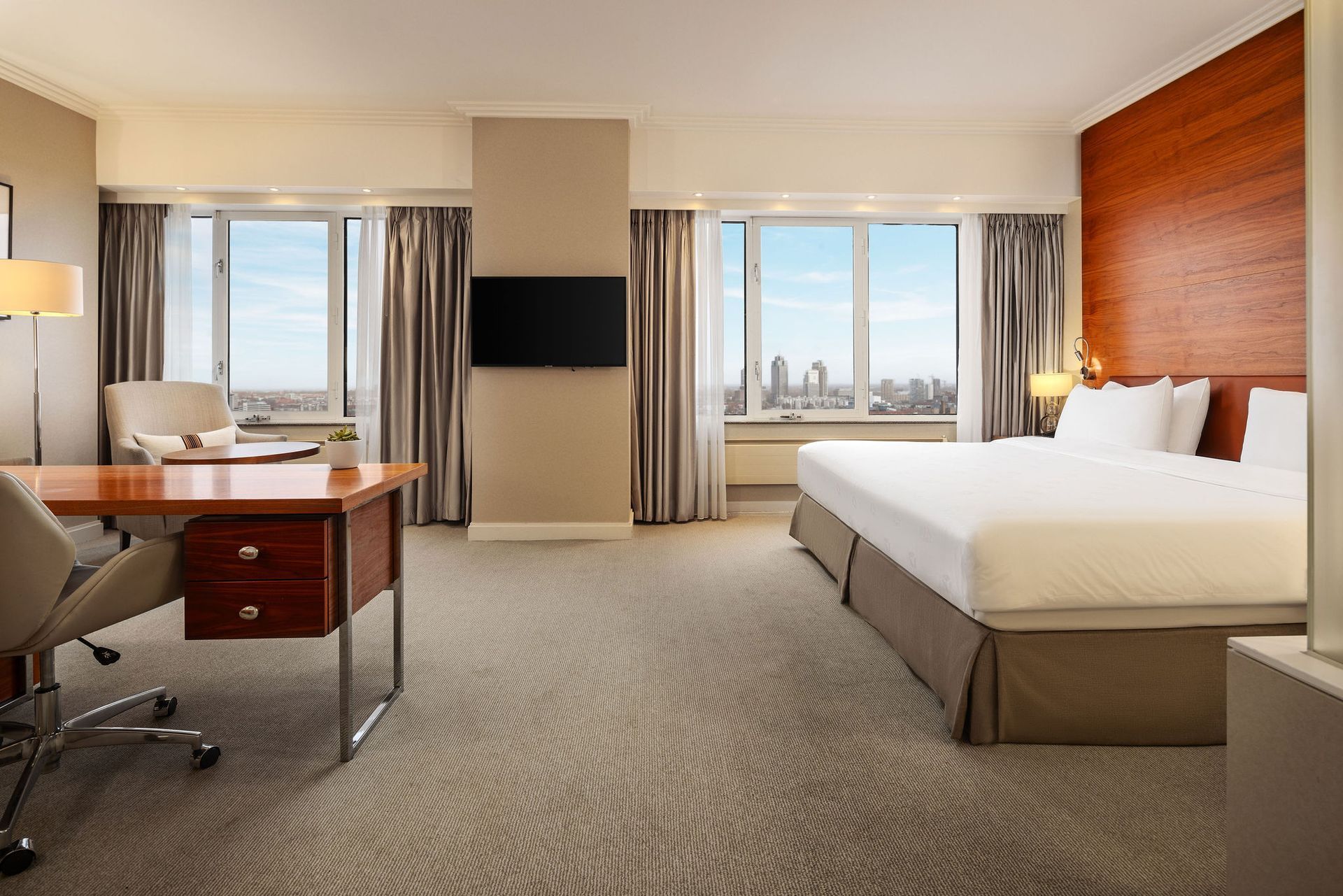 Hotel room with a bed, desk, and large windows with city view.