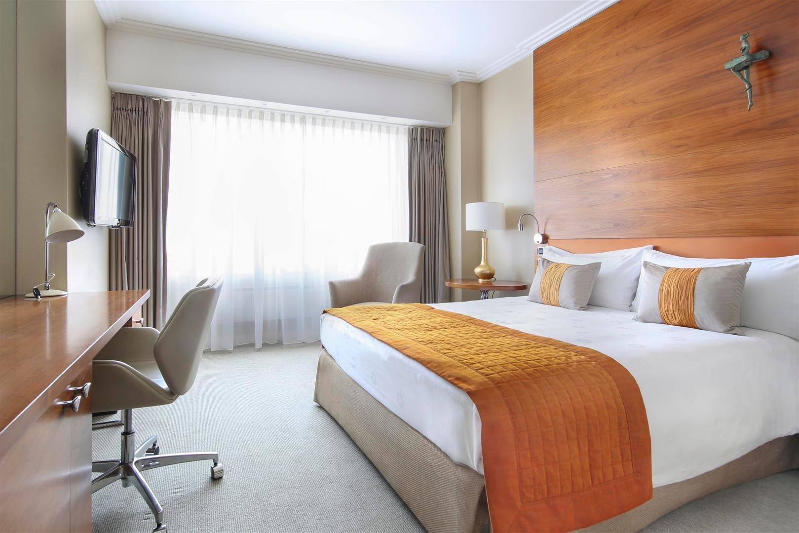 Hotel room with a bed, desk, and chair. Large wooden headboard, window with curtains, and neutral colors.