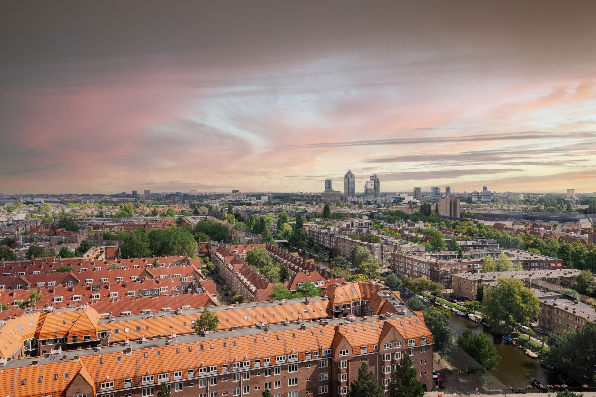 Cityscape view with rows of orange rooftops, green trees, and buildings under a cloudy sky with pastel colors.