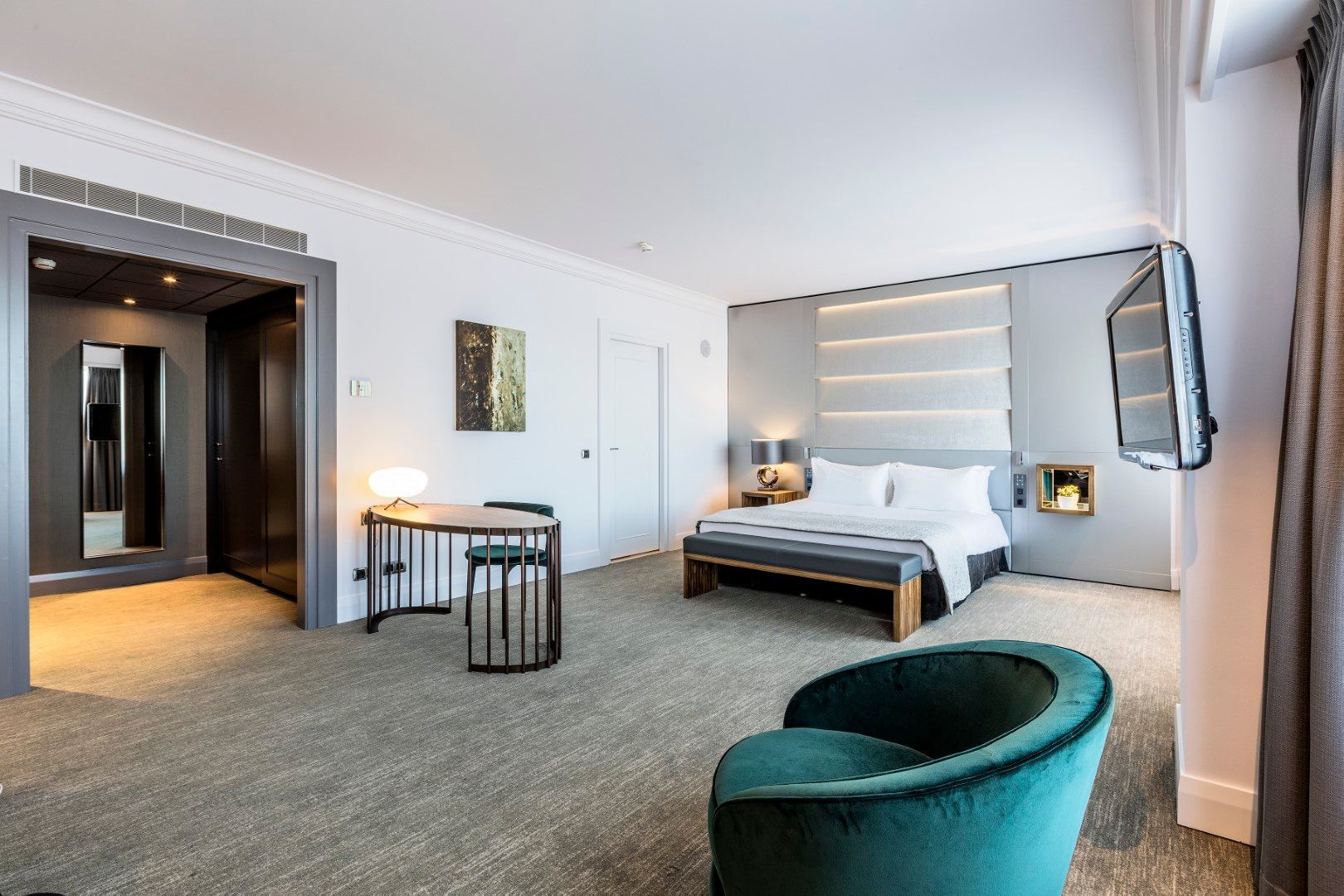 Hotel bedroom with bed, desk, chair, and TV; gray and teal color scheme.