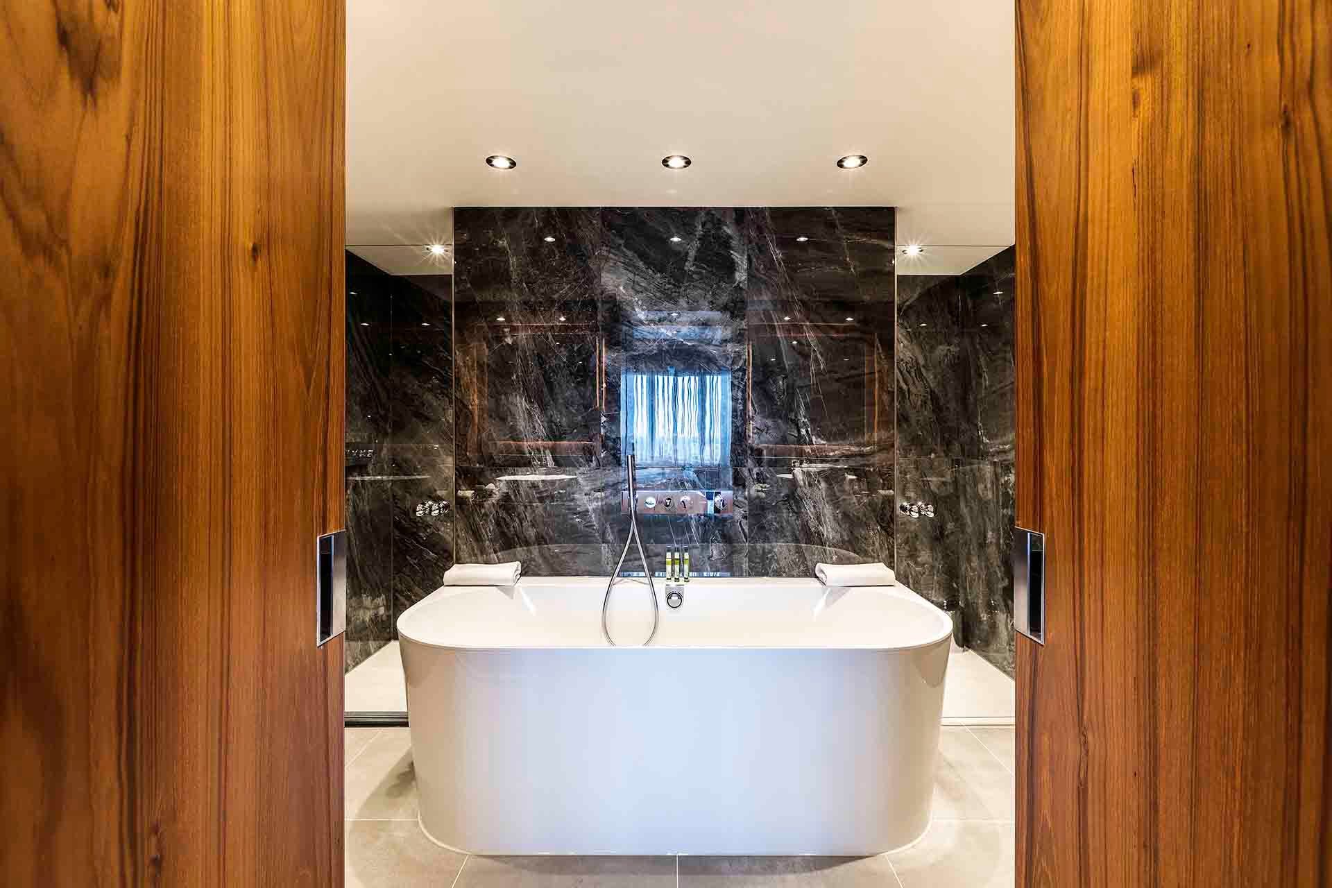 Luxurious bathroom with a white tub in front of a dark marble wall, framed by wooden doors.