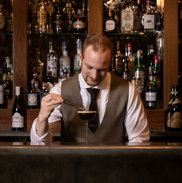 Bartender in vest garnishes a cocktail behind a bar with many bottles, using a tool.