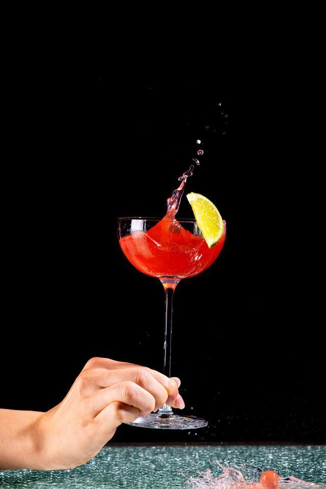 Cocktail in a glass with liquid splashing upwards, hand holding the stem, lime wedge, black background.
