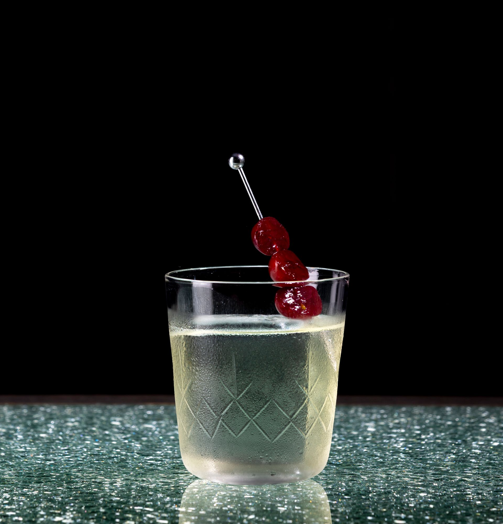 Cocktail in a cut-glass tumbler, garnished with cherries on a skewer, against a dark background.
