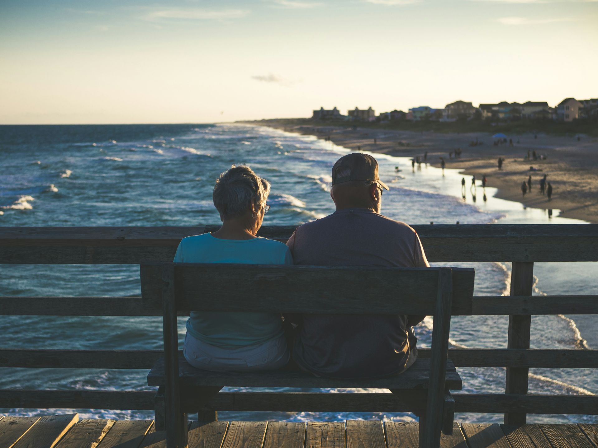 Elderly couple seated on a pier bench, looking at the ocean and beach at sunset.