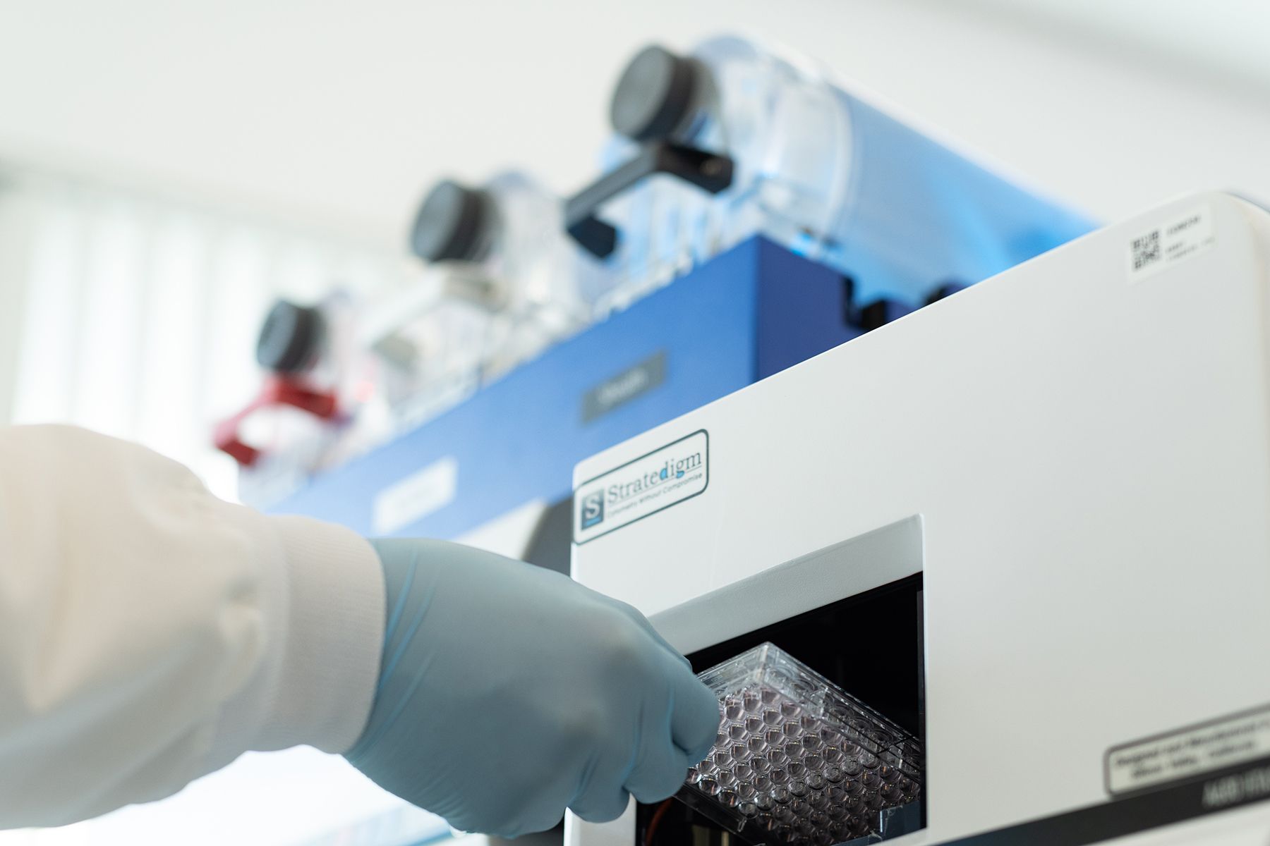 Person in blue glove inserts a multi-well plate into a laboratory machine, with other equipment visible.