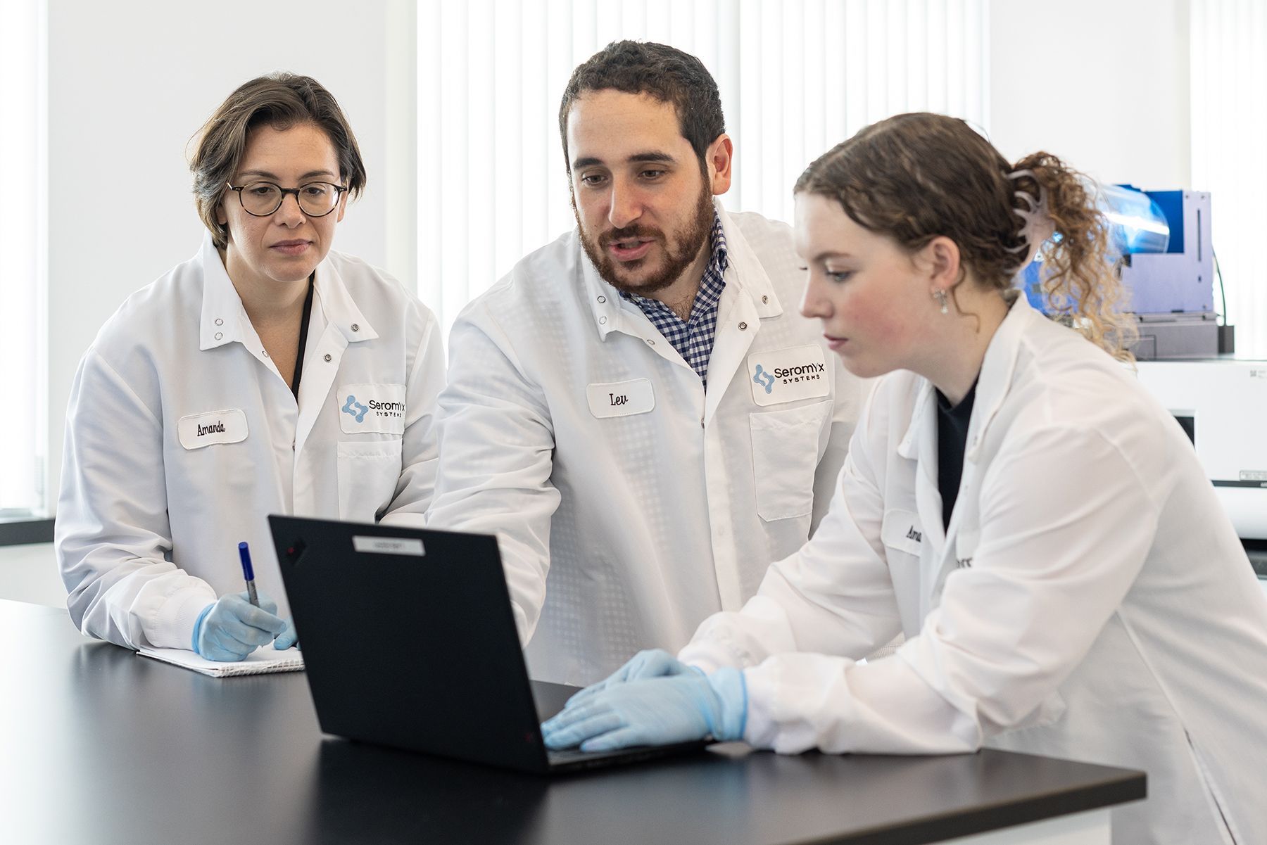 Three scientists in lab coats reviewing data on a laptop, one writing notes in a laboratory setting.