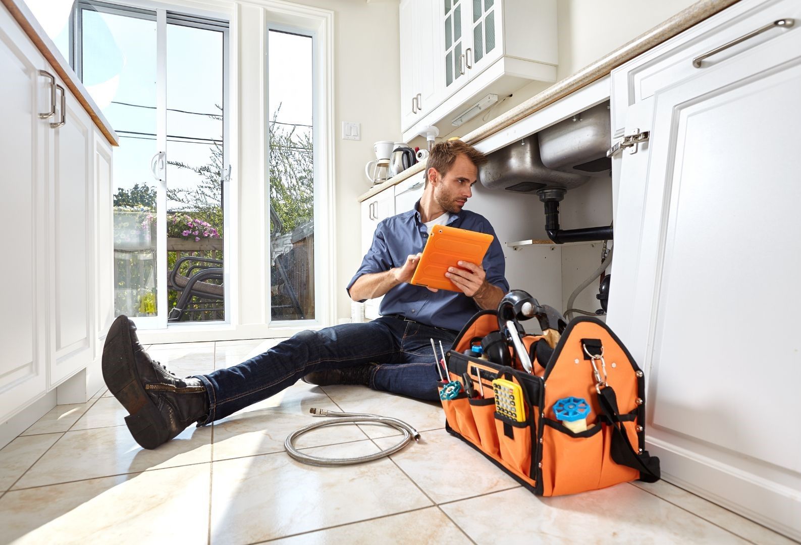 A plumber is sitting on the floor in a kitchen looking at a tablet.