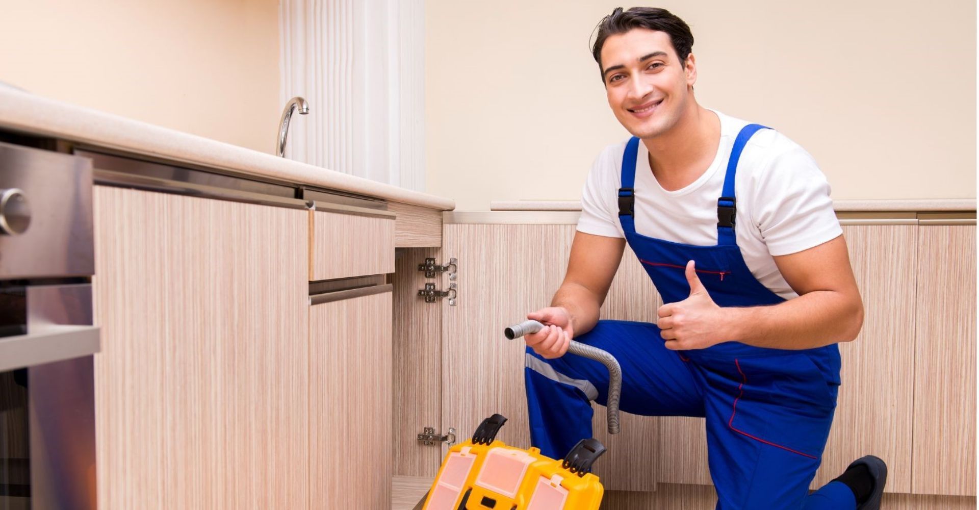 A plumber is fixing a sink in a kitchen and giving a thumbs up.