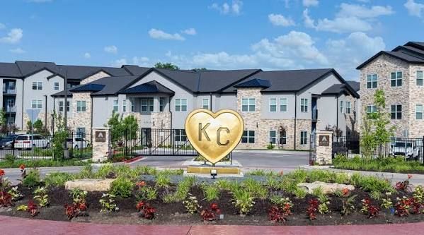 Apartment complex entrance with landscaped flowers and a gold KC heart sign under a blue sky
