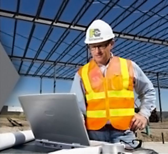 Construction worker in hard hat and orange vest using a laptop at a building site under a steel roof framework