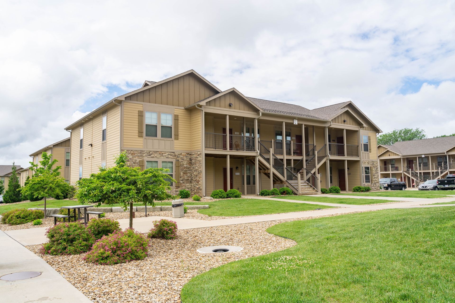 Apartment complex with stone and beige siding, balconies, and a landscaped lawn under a cloudy sky
