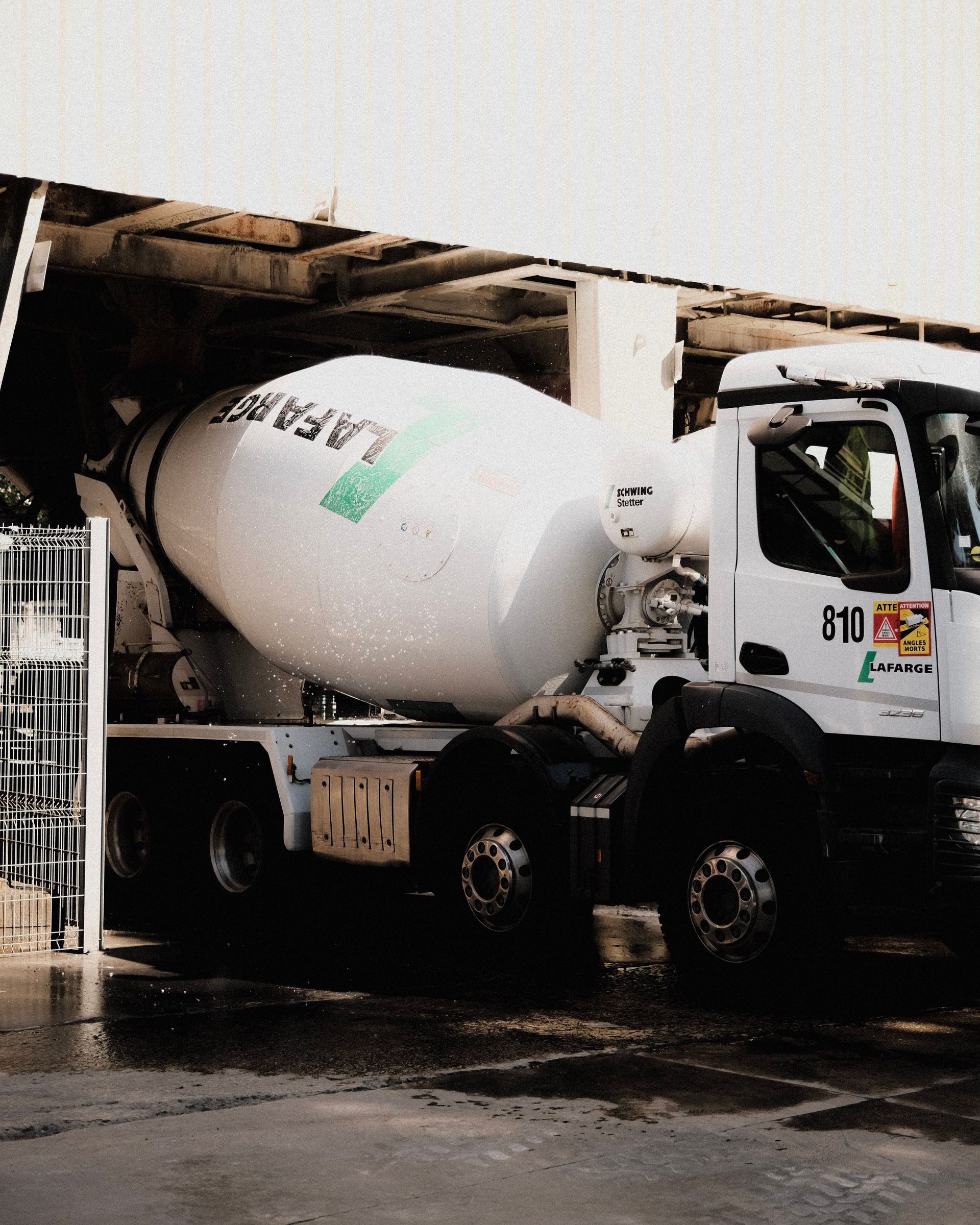 Cement mixer truck with a white drum drives through a wet industrial area under a covered structure