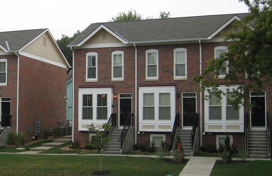 Row of brick townhouses with front steps, white trim, and small lawns along a sidewalk.