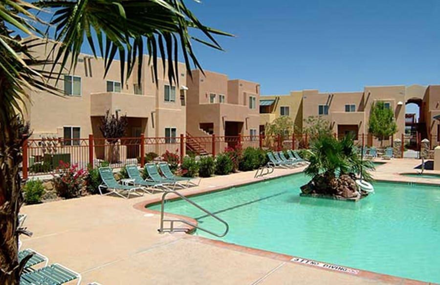 Desert resort pool with turquoise water, tan buildings, lounge chairs, and palm trees under a clear blue sky