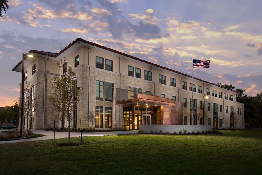 Modern stone office building at dusk with lit entrance, lawn, and American flag flying outside
