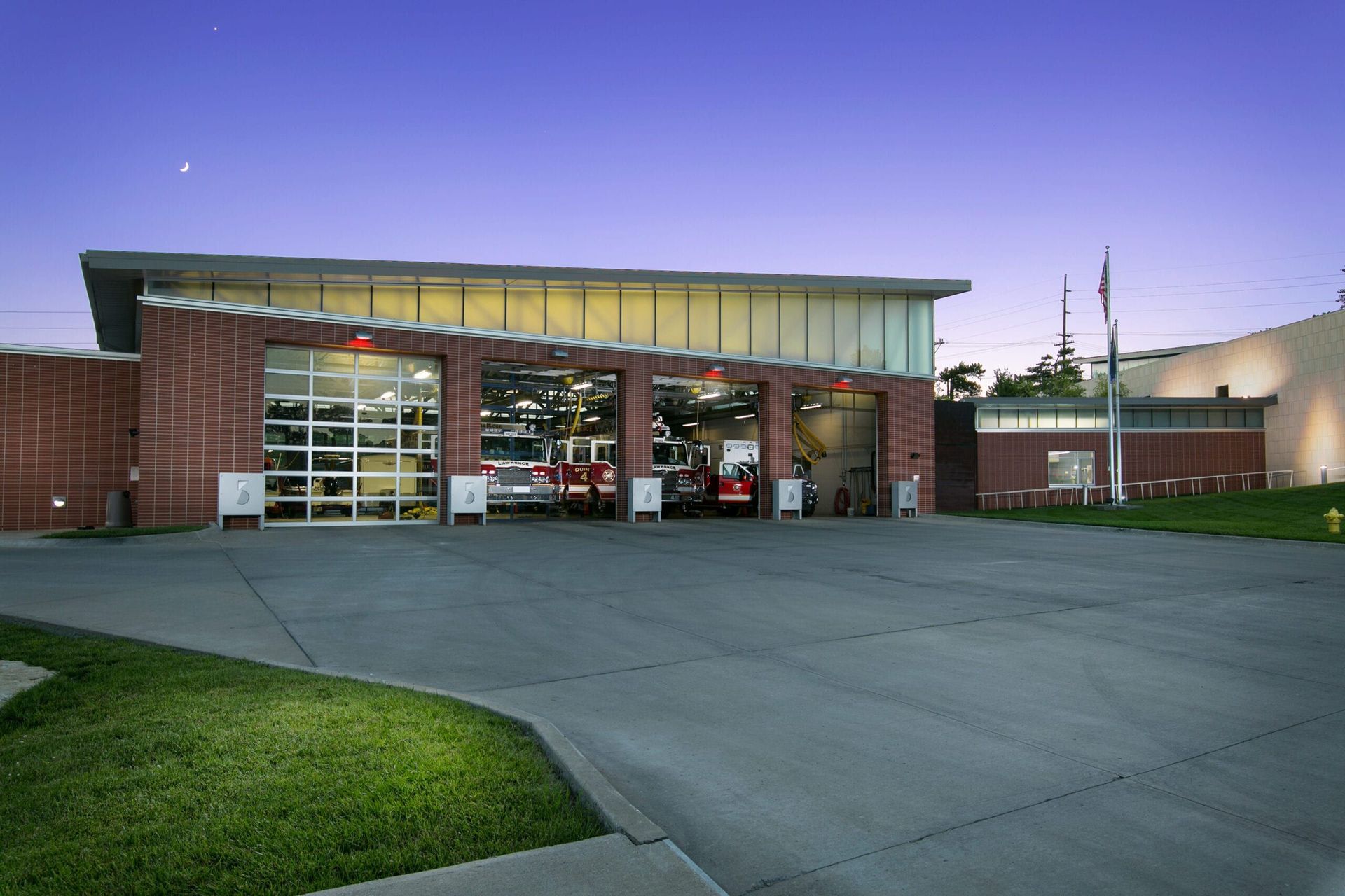 Fire station exterior at dusk with open garage bays and fire trucks inside