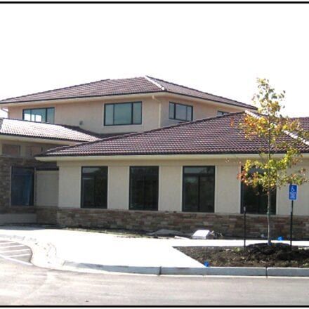 Two-story beige building with a red tile roof, dark windows, and a parking area in front