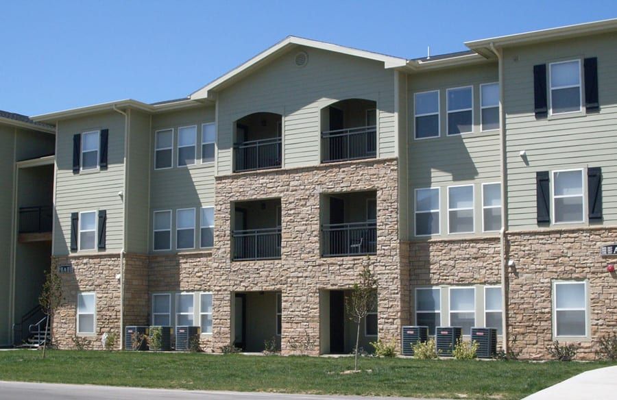 Three-story apartment building with tan siding and stone accents under a clear sky