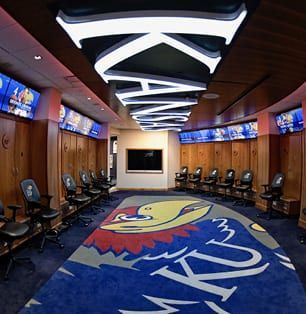 Modern locker room with blue carpet, wood lockers, black chairs, and illuminated ceiling panels