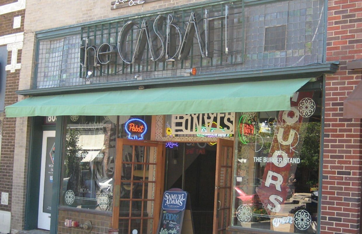 Storefront with a green awning, wooden door, and neon open sign on a brick street facade