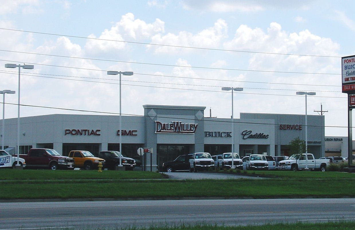 Car dealership lot with parked cars and showroom buildings beside a road under a cloudy sky