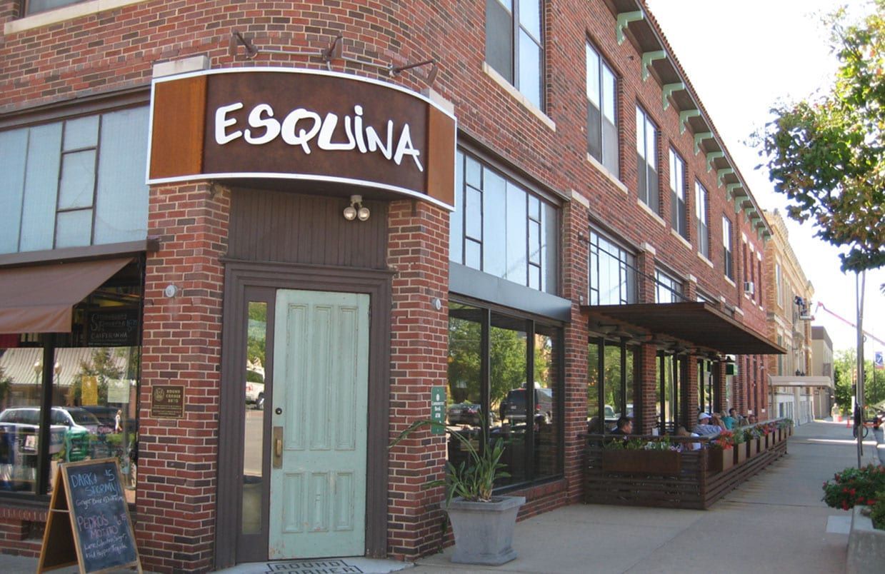 Brick corner storefront with a black awning reading “ESQUINA” and outdoor seating along the sidewalk.