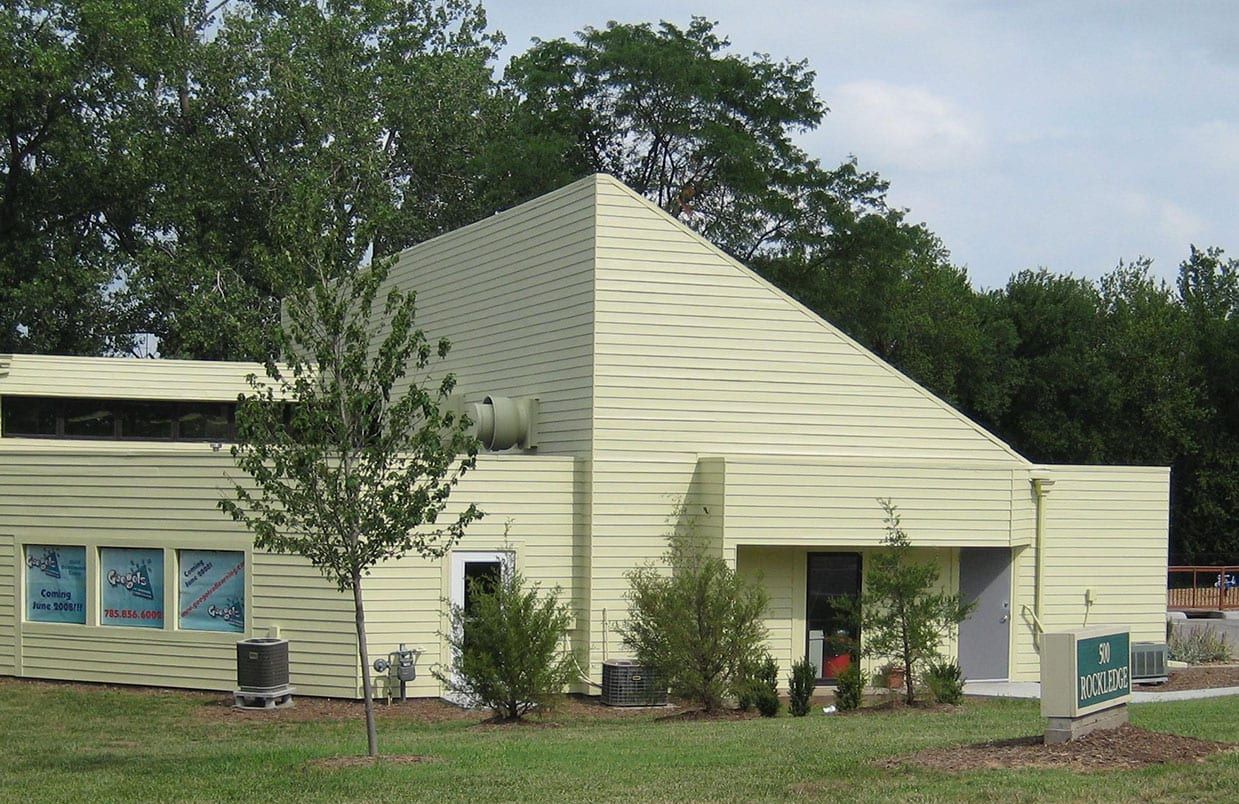 Small beige building with a sloped roof and trees in front, beside a grassy area