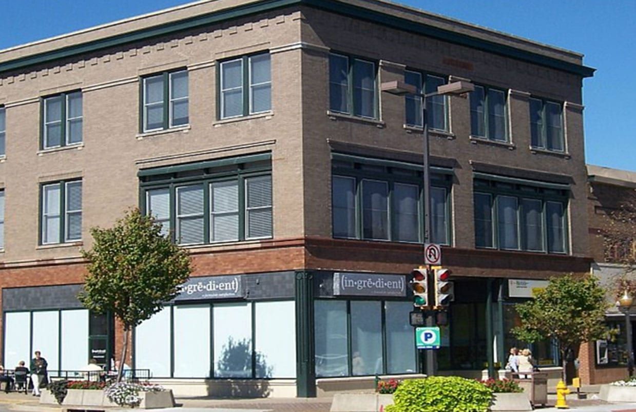 Brick corner building with large windows, storefronts, and a sidewalk in front