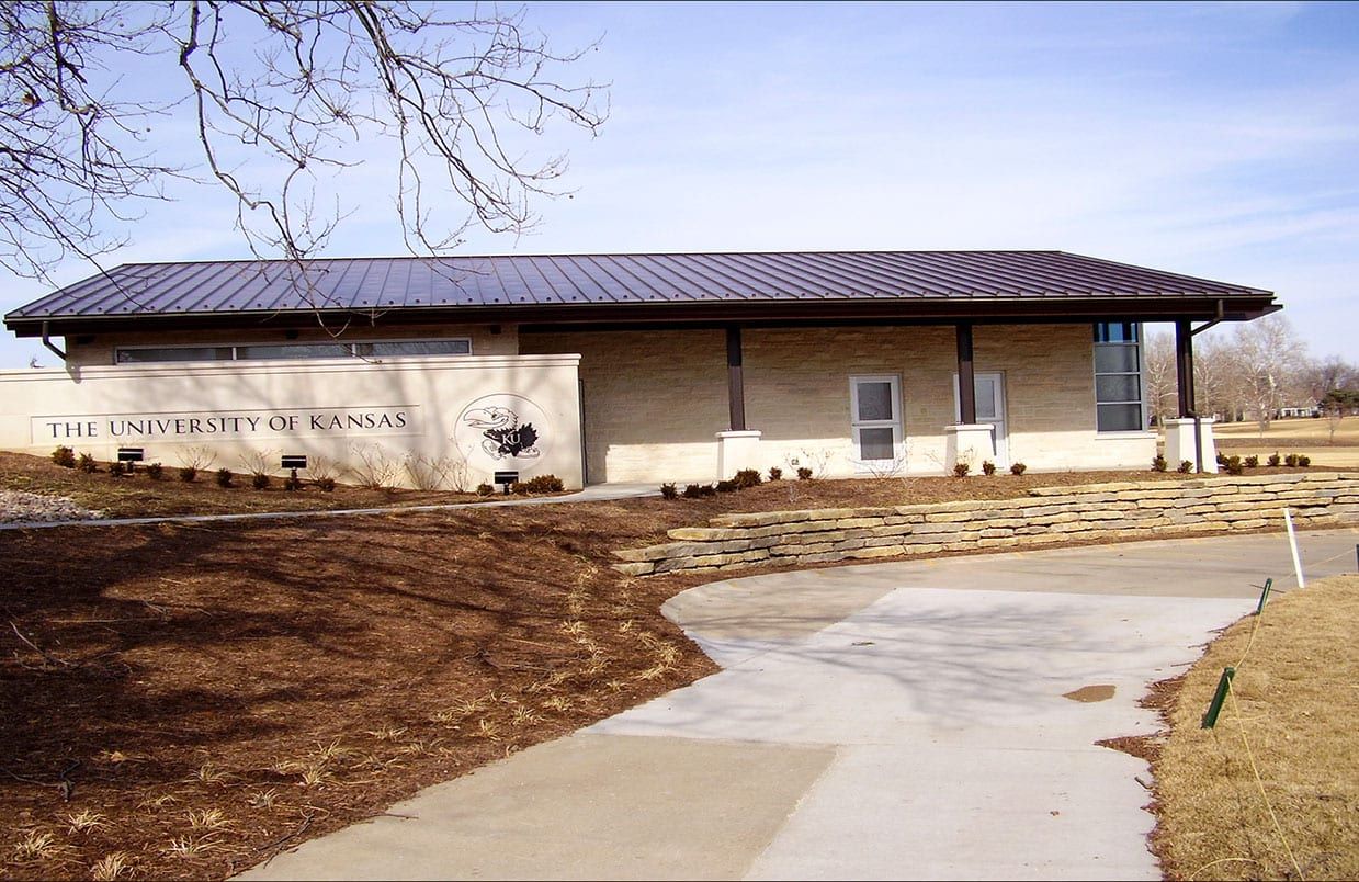 Single-story beige building with dark roof and a curved concrete walkway in a landscaped park setting