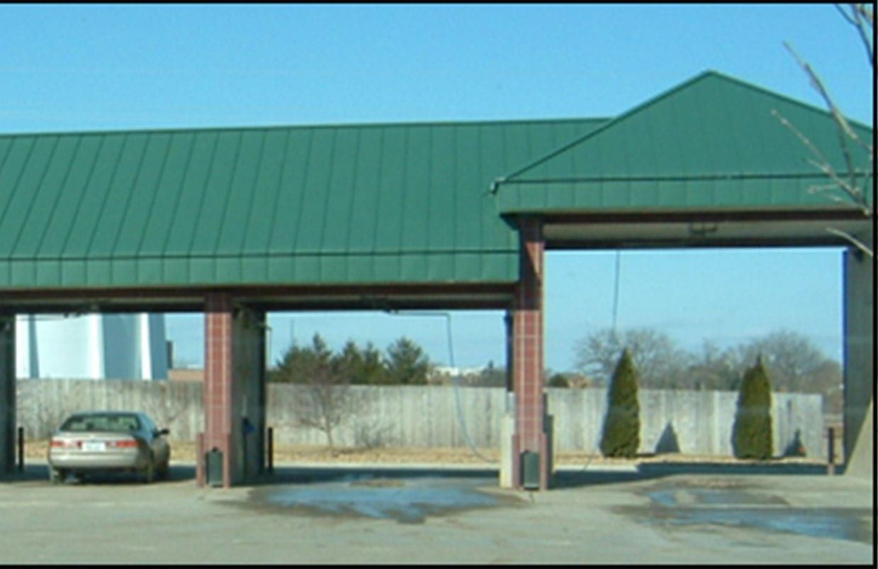 Green-roofed carport with a parked sedan and a few trees in the background.
